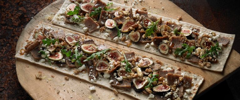 Two flatbreads on a pizza paddle that are stuffed with gyro meat slices, fresh figs, feta cheese, nuts and arugala